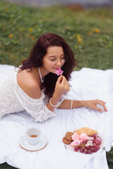 A young woman in a Korean fashion-inspired summer outfit lies on a blanket, holding a flower during a picnic with tea and pastries. The serene and stylish setting captures themes of relaxation, beauty