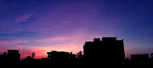 Obraz premium Silhouetted buildings stand against a vibrant sunset sky transitioning from pink to deep blue. The urban skyline is outlined in shadow, with a few palm trees visible amongst the structures.
