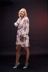 A woman in a dress smiles and poses for the camera. The dress is white and has a floral pattern. The woman is standing against a dark background, which creates a contrast.