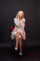 A woman in a dress smiles and poses for the camera. The dress is white and has a floral pattern. The woman is standing against a dark background, which creates a contrast.