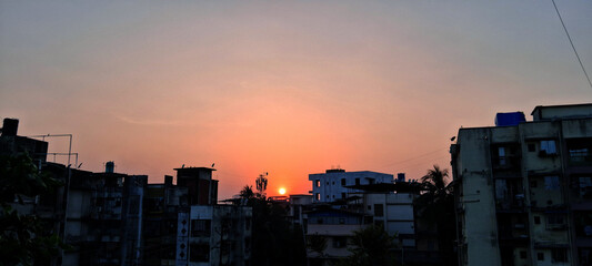 A vibrant sunset casts a gradient of colors from orange to deep blue over the skyline. Silhouetted buildings stand in contrast against the colorful sky, creating a peaceful urban landscape.
