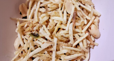 A close-up view of a bowl filled with crispy, straw-shaped snacks mixed with peanuts and herbs. The thin, golden sticks are scattered randomly, suggesting a savory and crunchy treat.
