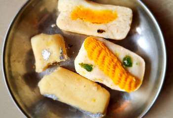 Various pieces of Indian sweets are arranged on a metal plate, some adorned with silver leaf. The confections show a range of colors and textures, indicating different flavors and ingredients.