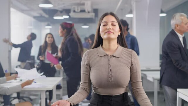A young modern corporate office woman sitting quietly in a yoga pose meditating with closed eyes unaffected by the chaos, commotion, fighting, mess created by the colleagues or coworkers at workplace