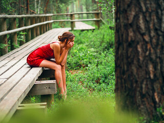 Young teenager girl in high fashion red dress posing in a forest park sitting on a wooden footpath, she has sad look. Prom photo shoot. Model short hair and slim body type. Stress concept.