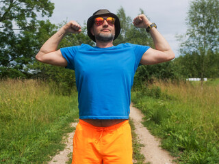 Naklejka premium A man in a blue shirt and orange shorts flexes his hands. The man is wearing a hat and he is enjoying the outdoors. Male in his 40s with grey beard. Sport and fitness outdoor.