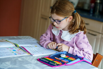 Little girl with glasses, wearing a pink sweater, sits at a table doing homework. Child looks up...