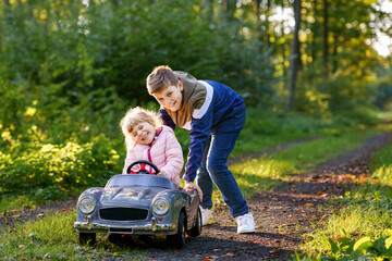 Two happy children playing with big old toy car in autumn forest, outdoors. Kid boy pushing and driving car with little toddler girl, cute sister inside. Laughing and smiling kids. Lovely family