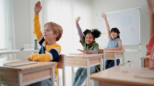 Elementary school students are sitting in the classroom at school and are raising their hands when the teacher asks