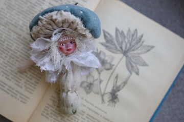 Unique doll resembling a mushroom placed on a vintage book with floral illustrations at home. A whimsical doll shaped like a mushroom with delicate features rests atop an open vintage book