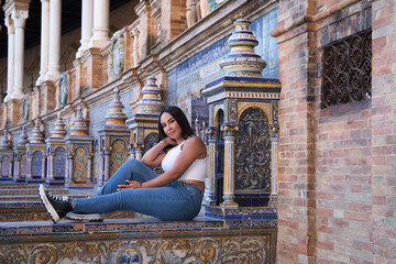 Young, pretty, brunette moroccan woman dressed in white top and jeans sitting on a tiled bench in the square of Spain in Seville. The woman is happy and smiling. Travel and tourism concept.