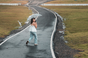 Fototapeta premium A young girl walks alone on a rainy road with an umbrella, embracing solitude and nature's beauty along her journey