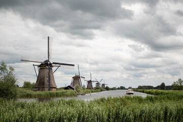 Group of historic windmills at the river with a passenger boat four tourists. Bushes and reed grass lines the shore. Kinderdijk in Netherlands, cloudy day.