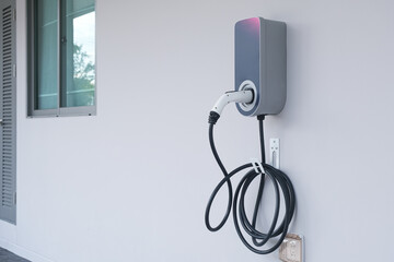 Modern electric vehicle (Ev) charging station with power cable plug for charging Ev or hybrid car, isolated on white house wall.