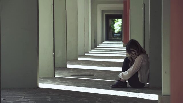 A woman is sitting on the ground under the portico of a building, curled up on herself, looking sad and alone, reflecting her feelings of isolation.
