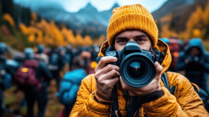 Obraz premium A candid shot of a photographer in a bright yellow beanie, smiling while holding a camera, surrounded by people in a picturesque outdoor setting filled with autumn colors.