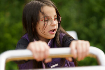 A little cute smiling girl in glasses and a purple vest plays on the playground. Happy childhood...
