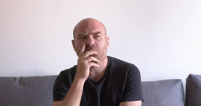 Bald man with a red goatee sitting on the couch, thinking and looking around with his eyes, searching for an idea, deep in thought.