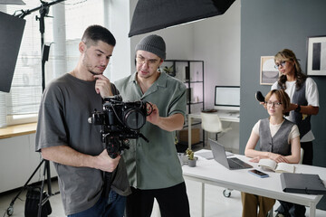 Young adult cameraman and director looking at camera display while discussing footage while make up artists brushing actress