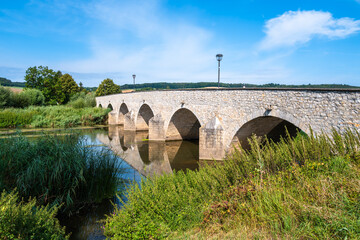 Fototapeta premium Stone bridge with arches over Wornitz river in Germany, surrounded by lush greenery.