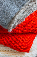 Grey, white and bright orange knitted sweaters in a pile. Stack of cozy handmade clothes. Different knitting patterns concept. Colourful woven jumpers. Knitted comfortable clothing. Knitted fashion.