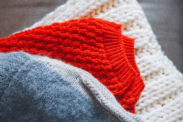 Grey, white and bright orange knitted sweaters in a pile. Stack of cozy handmade clothes. Different knitting patterns concept. Colourful woven jumpers. Knitted comfortable clothing. Knitted fashion.