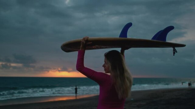 Young woman surfer walks along sand ocean beach, holds surfboard above head after sup training. Attractive girl walking or run to surfing with board. Evening dusk on bali, indonesia. Pink swuimsuit