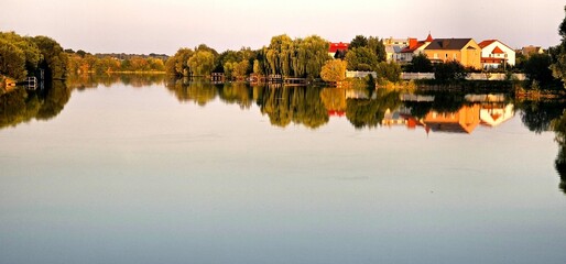 Calm waters reflect houses and greenery by a serene lake during golden hour in a peaceful countryside setting