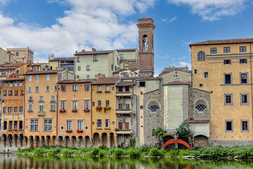 Colorful medieval houses sit above the Arno River in Florence, showcasing vibrant facades and historical architecture under blue cloudy sky.