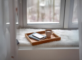 Cozy window seat scene with a wooden tray holding a warm drink, smartphone, and notebook, resting on a fluffy surface.  Wintery view outside.