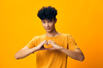 Young man with curly hair making a heart shape with hands on a vibrant yellow background, conveying...