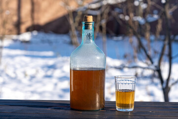 Big bottle with a drink made from fermented birch sap on the wooden table on winter sunny day, closeup. Traditional Ukrainian cold barley drink kvass in a glass jar and glass in yard