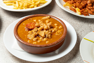 Close-up of traditional Spanish chickpea stew with chorizo, pork, and rich broth, served in a rustic clay pot on a white plate