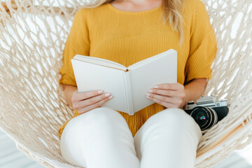Obraz premium A woman in yellow sweater sits comfortably in hammock, holding blank book while camera rests beside her. This serene scene captures moment of relaxation and creativity