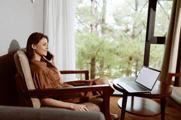 Happy woman in casual brown outfit talking on the phone while sitting in a chair near a laptop, enjoying a peaceful moment in a cozy interior with a green view