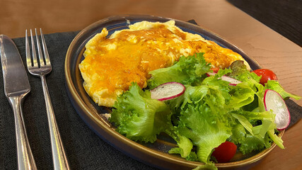 - A breakfast of a three-egg omelet with green salad leaves, radishes and tomatoes in a black ceramic plate with fork and knife stands on a brown wooden table.
