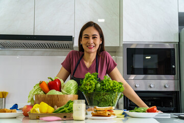 A beautiful Asian woman is standing behind island  of a modern kitchen with fresh vegetables and fruits lined up in front, with a cheerful expression on her face.