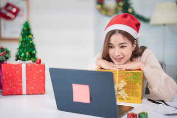 Smiling young asian businesswoman wearing santa hat, using laptop with christmas gift on desk in office during christmas holiday season, celebrating christmas and new year