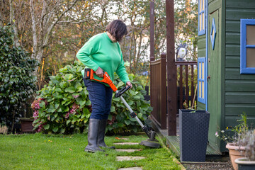 Woman Wearing Rubber Wellington Boots Using a Weed Whacker Strimmer along Lawn Edge and Stepping Stones