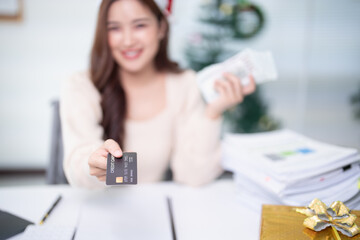 Happy businesswoman holding credit card and cash while making online christmas purchases, surrounded by financial documents, gifts and christmas decorations