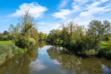  Wornitz river scene in Ebermergen with lush greenery on both banks, reflecting the blue sky and clouds. Trees with varying foliage surround the water
