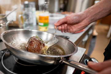 A close-up of a hand skillfully searing meat in a stainless steel pan on a stovetop. The rich,...