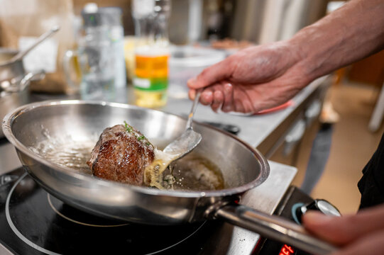 A close-up of a hand skillfully searing meat in a stainless steel pan on a stovetop. The rich, sizzling oil and herbs enhance the cooking process in a modern kitchen setting.