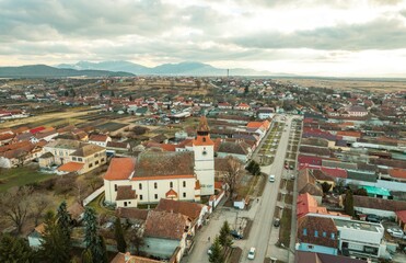 Naklejka premium Overhead view reveals the captivating landscape of Piatra Craiului and Rasnov, showcasing historic homes, sprawling fields, and distant mountains under a dramatic sky