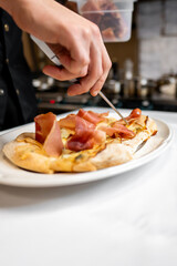 A chef adding slices of ham to a fresh, artisanal pizza on a white plate. The kitchen is softly blurred in the background, highlighting the delicious preparation process.
