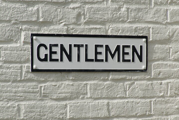 Sign for Gentlemen's toilet. Black lettering on white background on white brick wall.