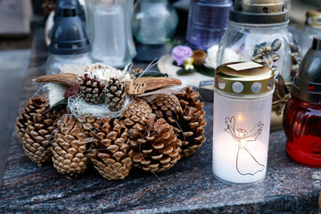 A decorative pine cone wreath with natural elements placed on a grave, accompanied by a glowing cemetery candle featuring an angel illustration. 