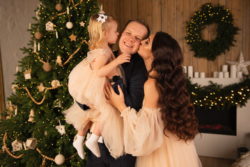 the wife and daughter kiss the husband against the background of a Christmas tree, a fireplace and Christmas paraphernalia