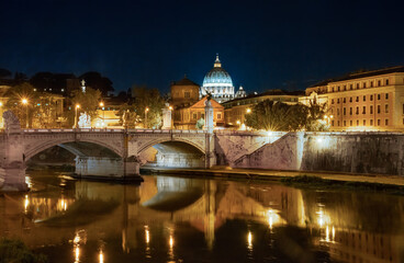 Seen from the Ponte Umberto (Umberto Bridge) across the Tiber River, the lighted iconic dome of St. Peter’s Basilica against the backdrop of Vatican City in Rome, Italy on a summer evening.