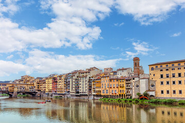 Colorful medieval houses sit above the Arno River in Florence, showcasing vibrant facades and historical architecture under blue cloudy sky.